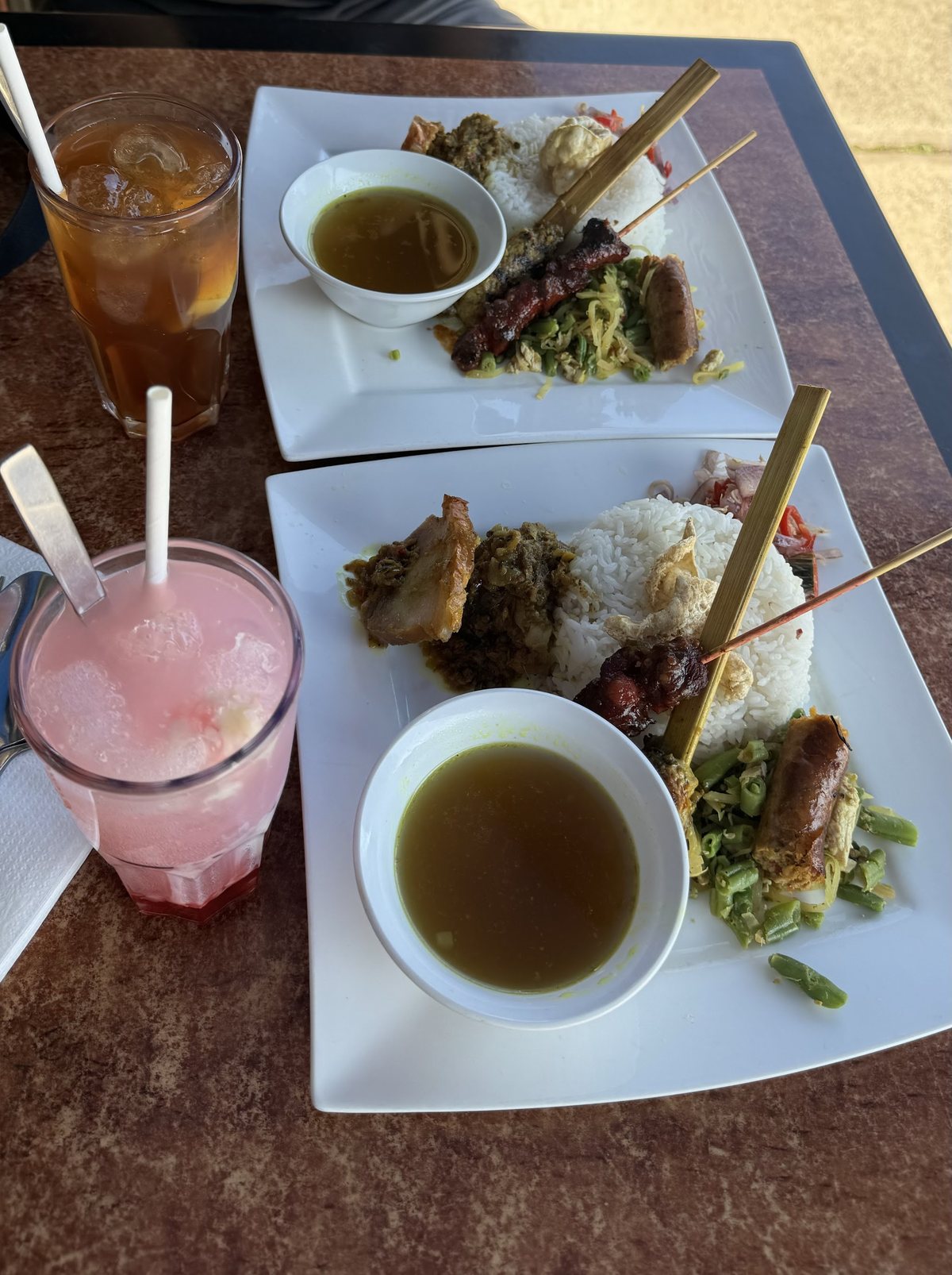 Two rice plates with satay, vegetables and soup, an iced tea and a pink drink at the front tables